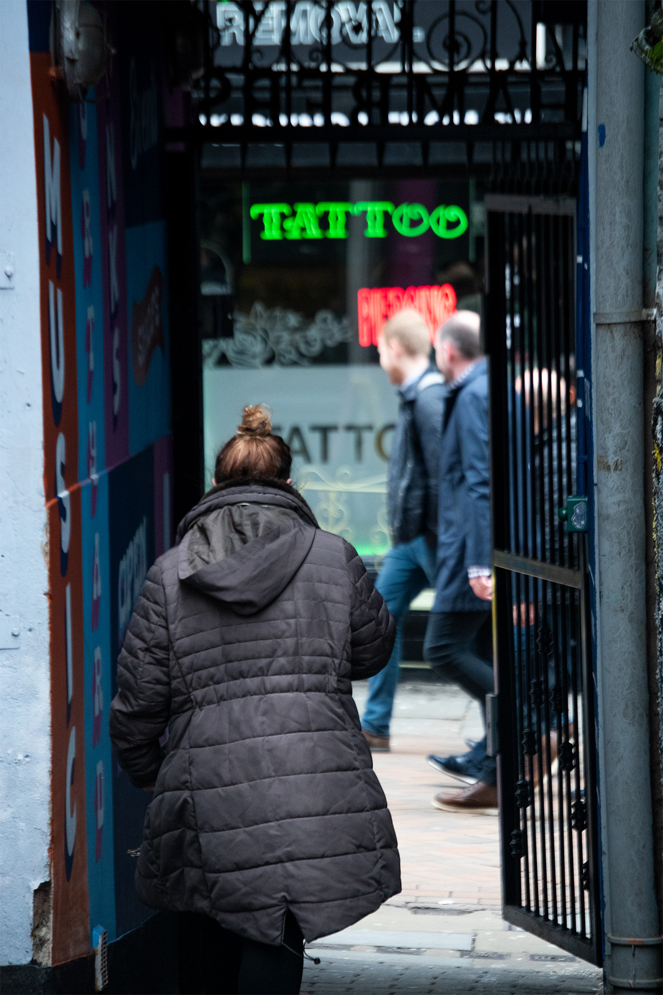 person walking into gated alleyway with neon signage for tattoo